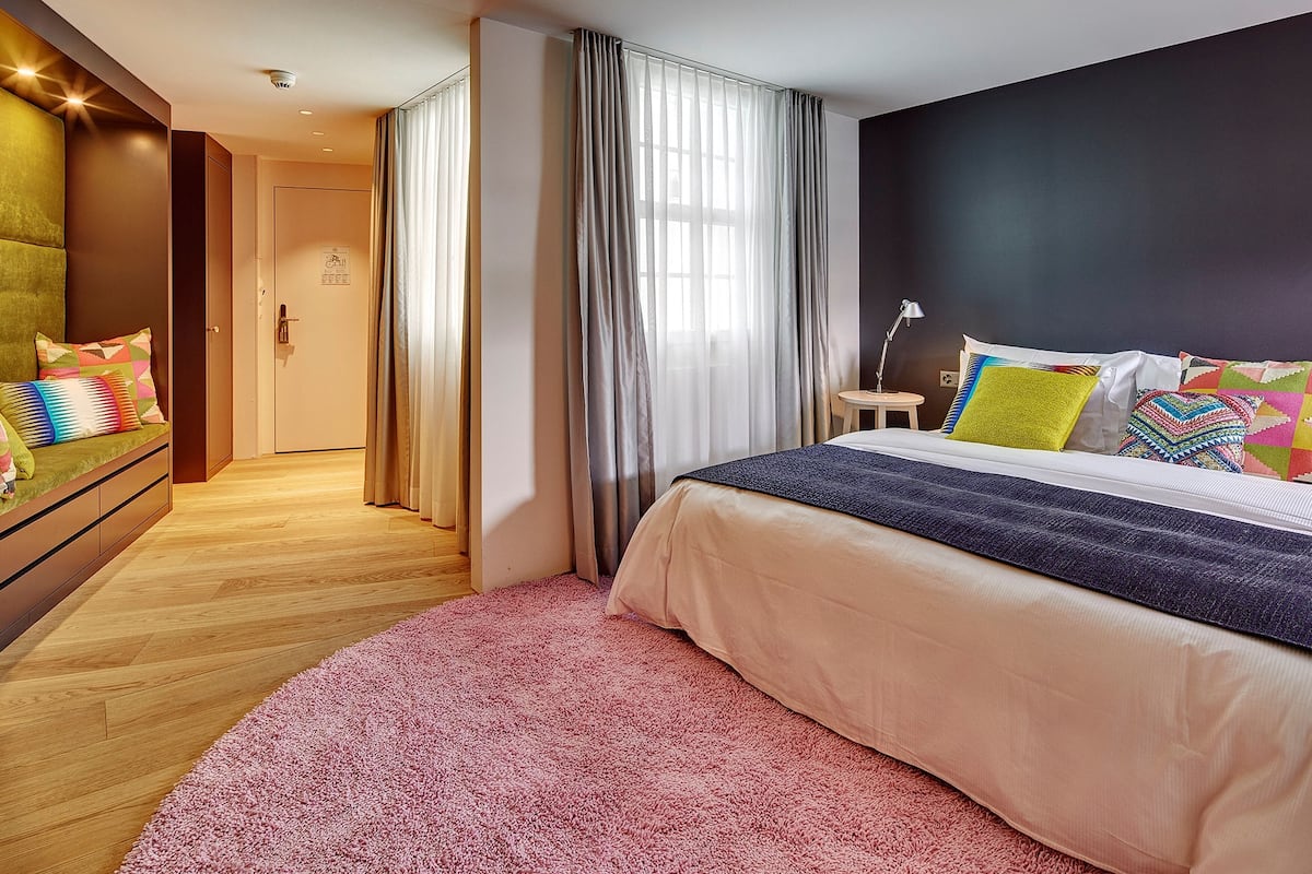 A modern hotel room with a large bed, a green sofa, a small table, and a view of the hallway.