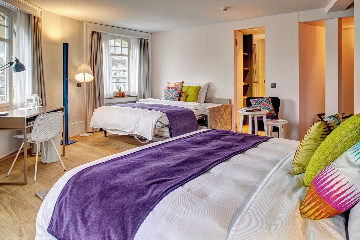 A hotel room with two beds, a desk, a chair, and a small table with a lamp.
