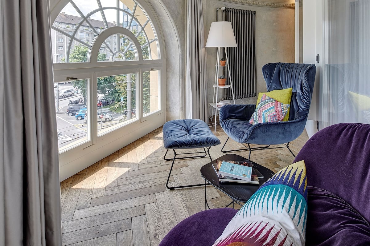 A modern living room with a large arched window, a blue armchair, a purple sofa, and a small black table with books.