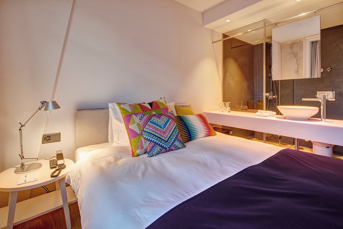 A modern hotel room with a bed, bedside table with a lamp, a colorful pillow, a bathroom with a sink and mirror.