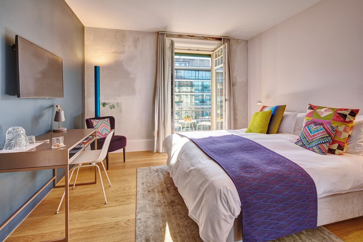 A modern hotel room with a bed, desk, chair, and a large window.