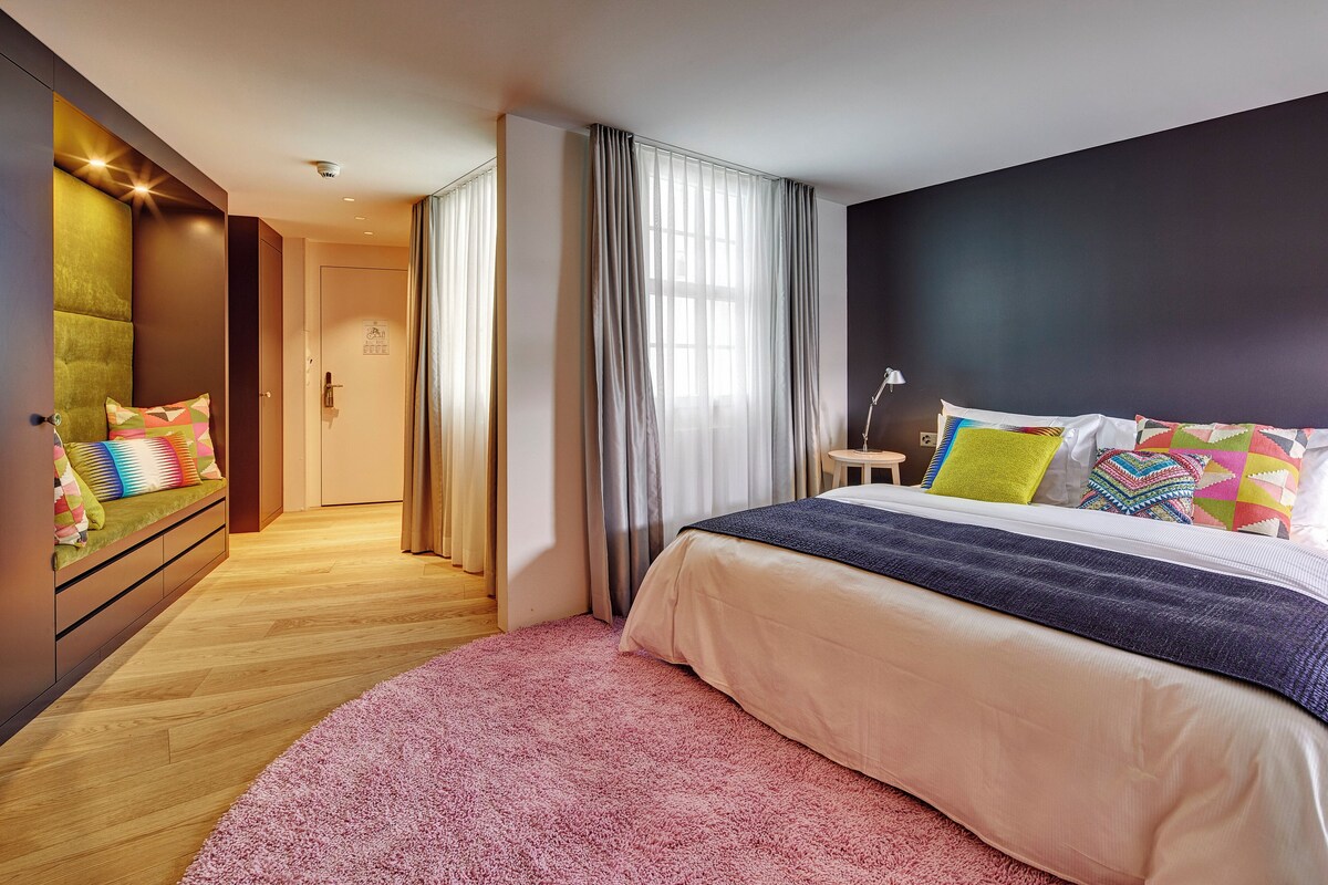 A bedroom with a large bed, a pink rug, a nightstand, and a wall-mounted TV.