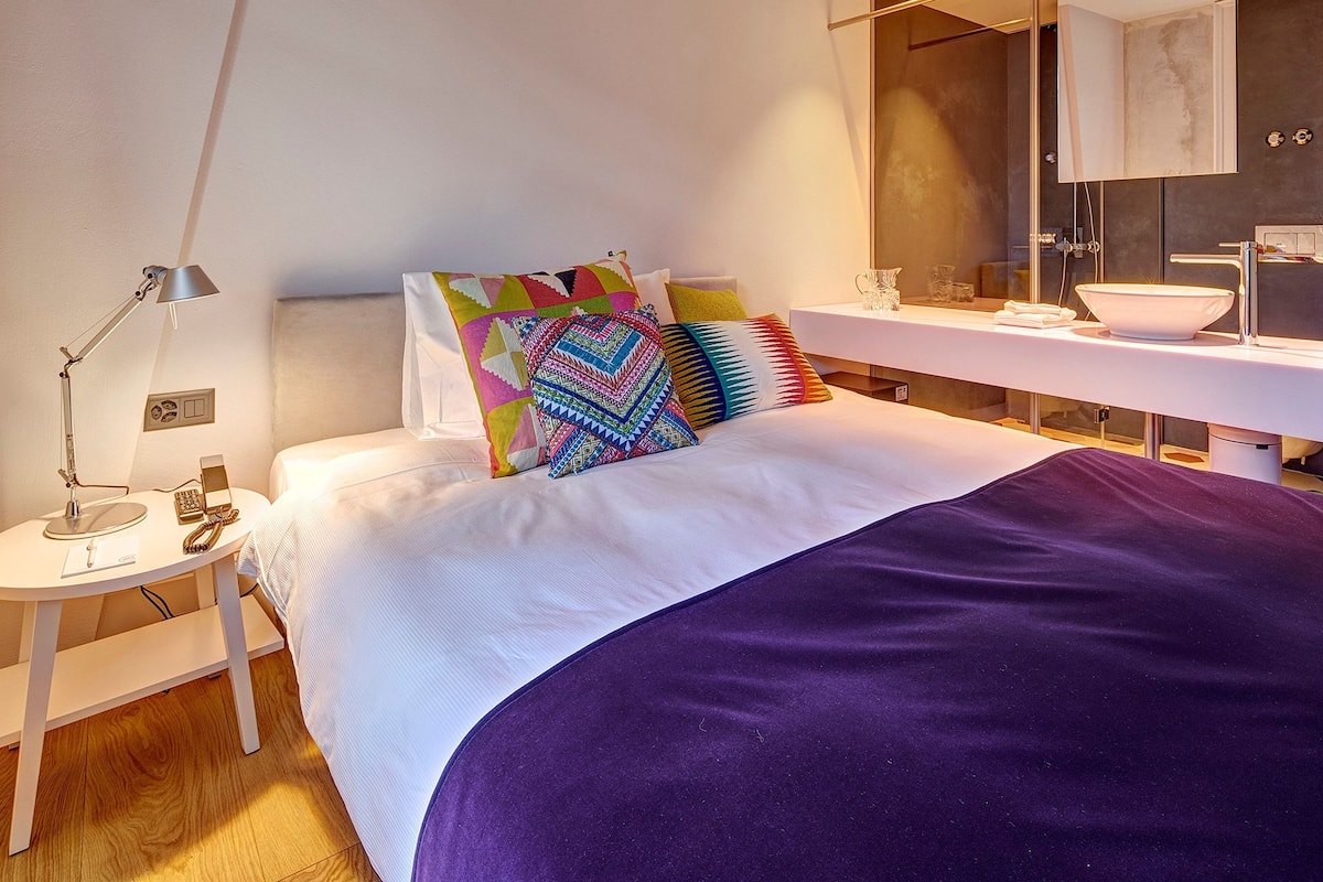 A modern hotel room with a neatly made bed, a bedside table with a lamp, a bathroom with a large mirror and a sink.