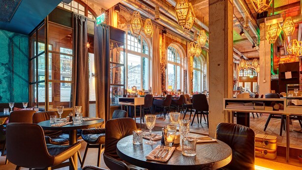 Un interior de restaurante con mesas redondas, sillas de cuero y vistas a la calle a través de grandes ventanales.