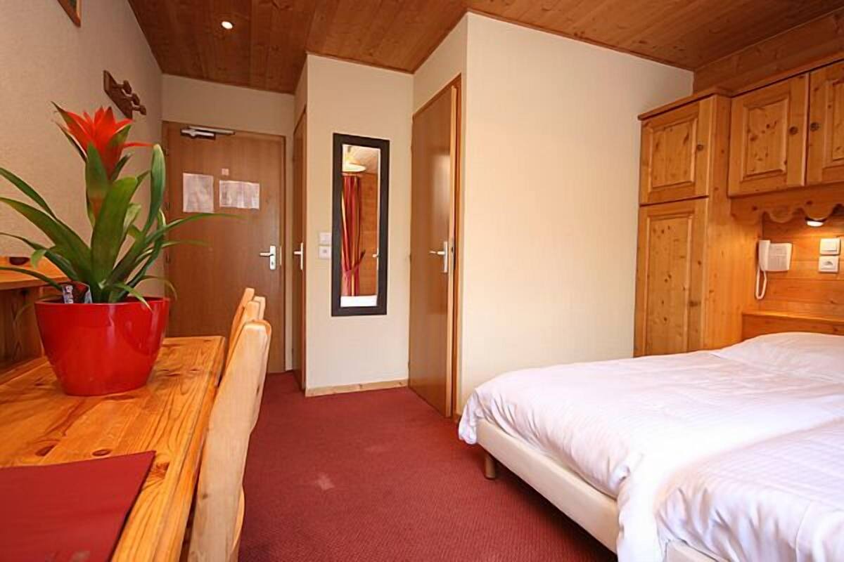 A hotel room with a wooden ceiling, a bed, a wooden cabinet, a red plant, and a red carpet.