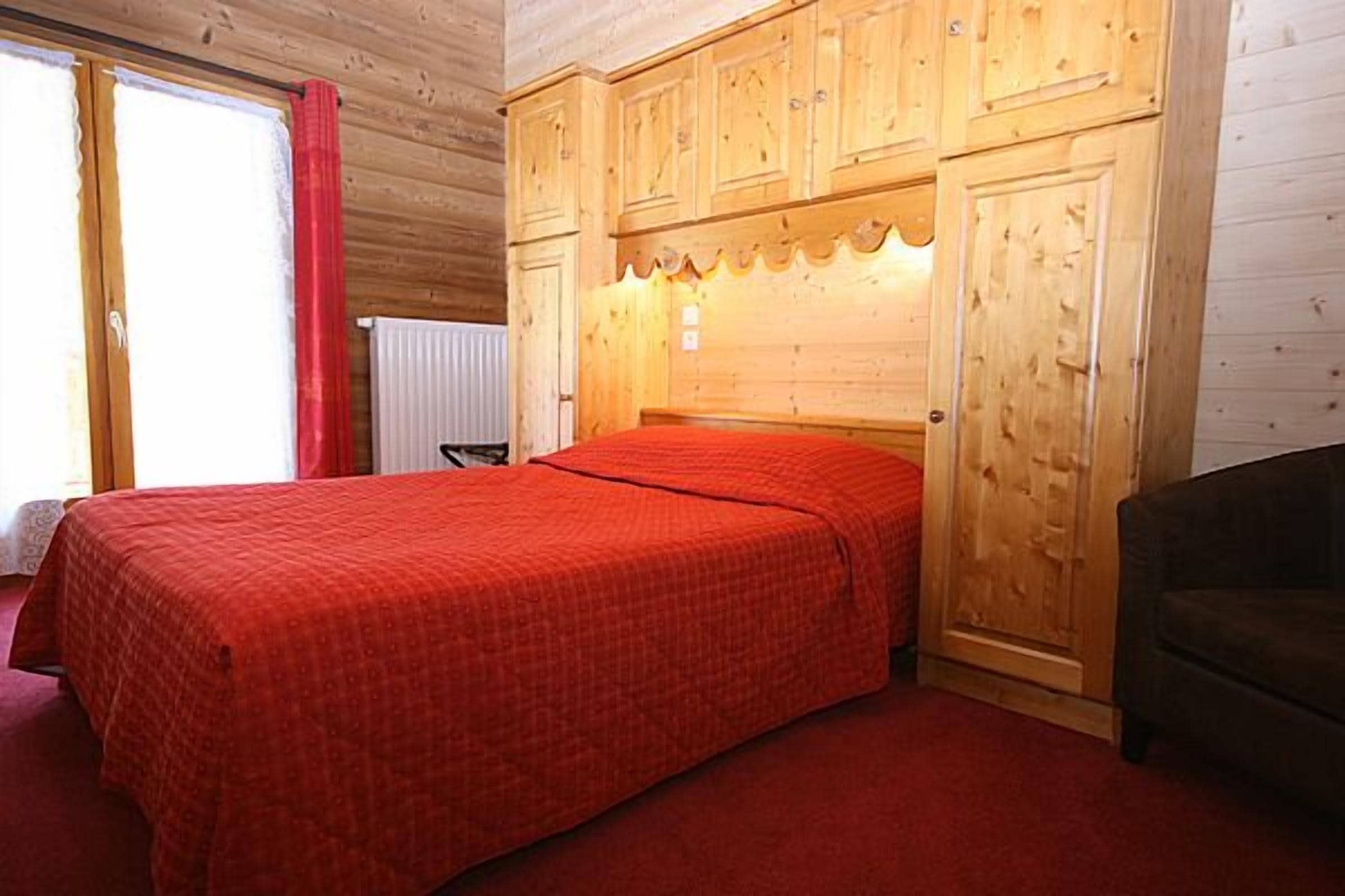 Ein Schlafzimmer mit einem hölzernen Kopfende, einer roten Bettdecke, einem schwarzen Stuhl und einem Fenster mit rotem Vorhang.