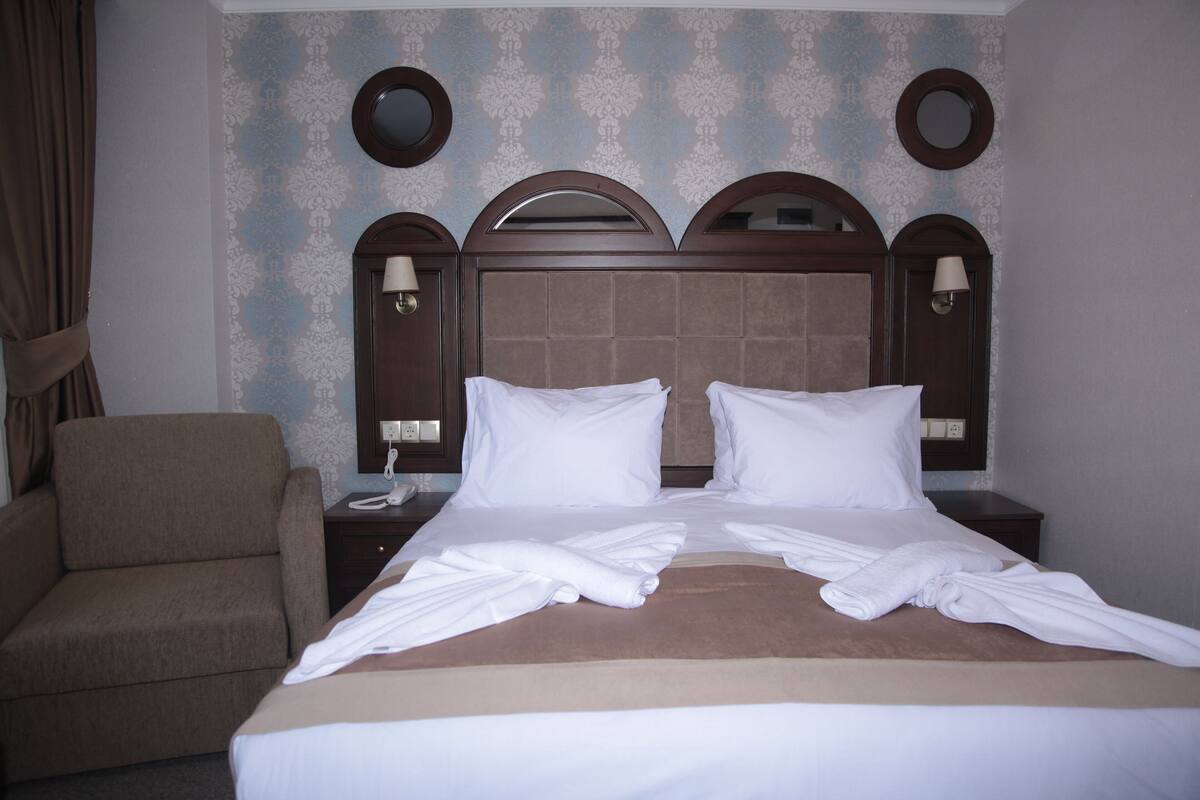 A hotel room with a large bed, two bedside tables, a chair, and a wall with decorative wallpaper and mirrors.