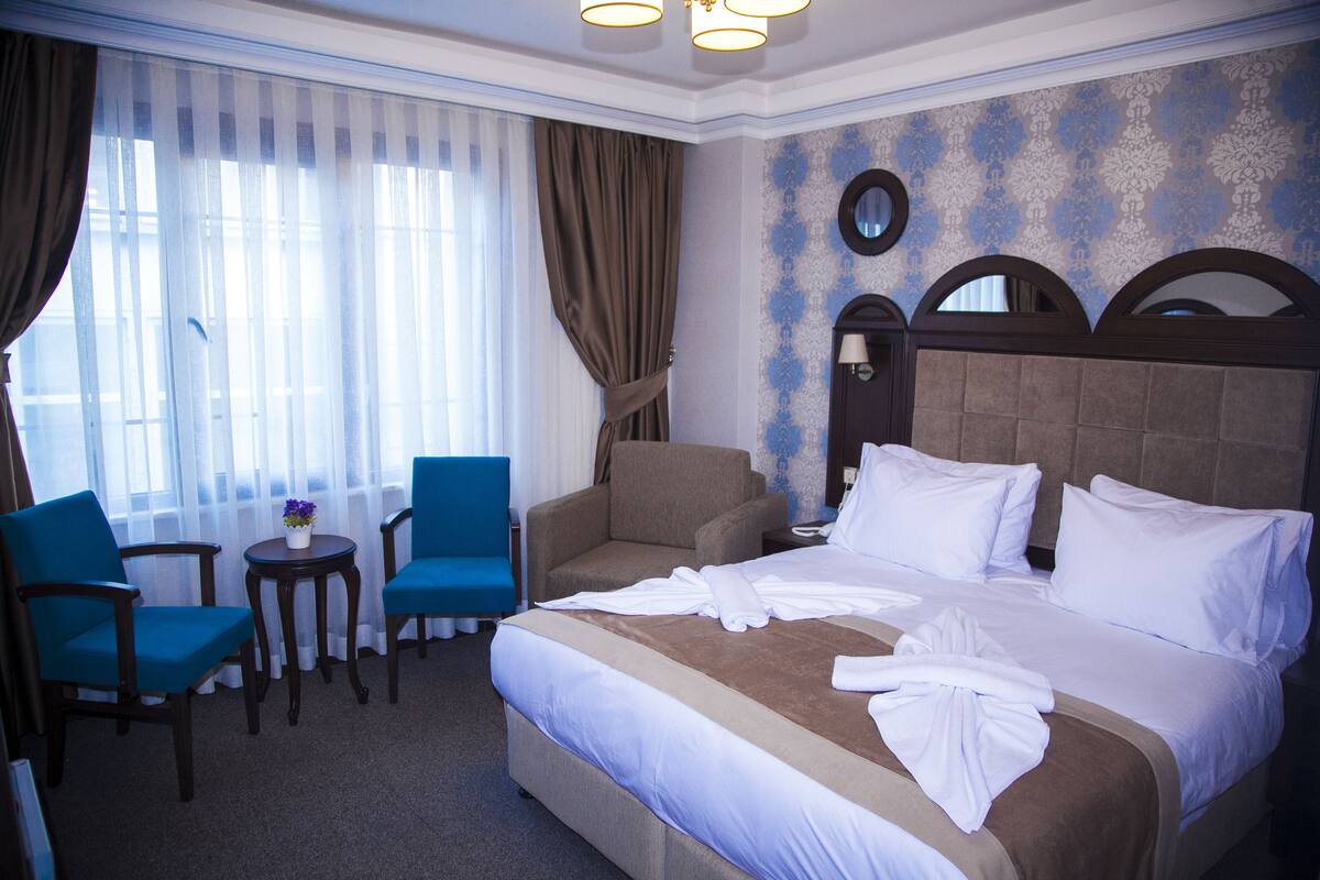 A hotel room with a large bed, two armchairs, a small table with a vase, and a window with curtains.