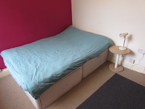 A small, single-bed room with a teal bedspread, a small round table with a lamp, and a red wall.