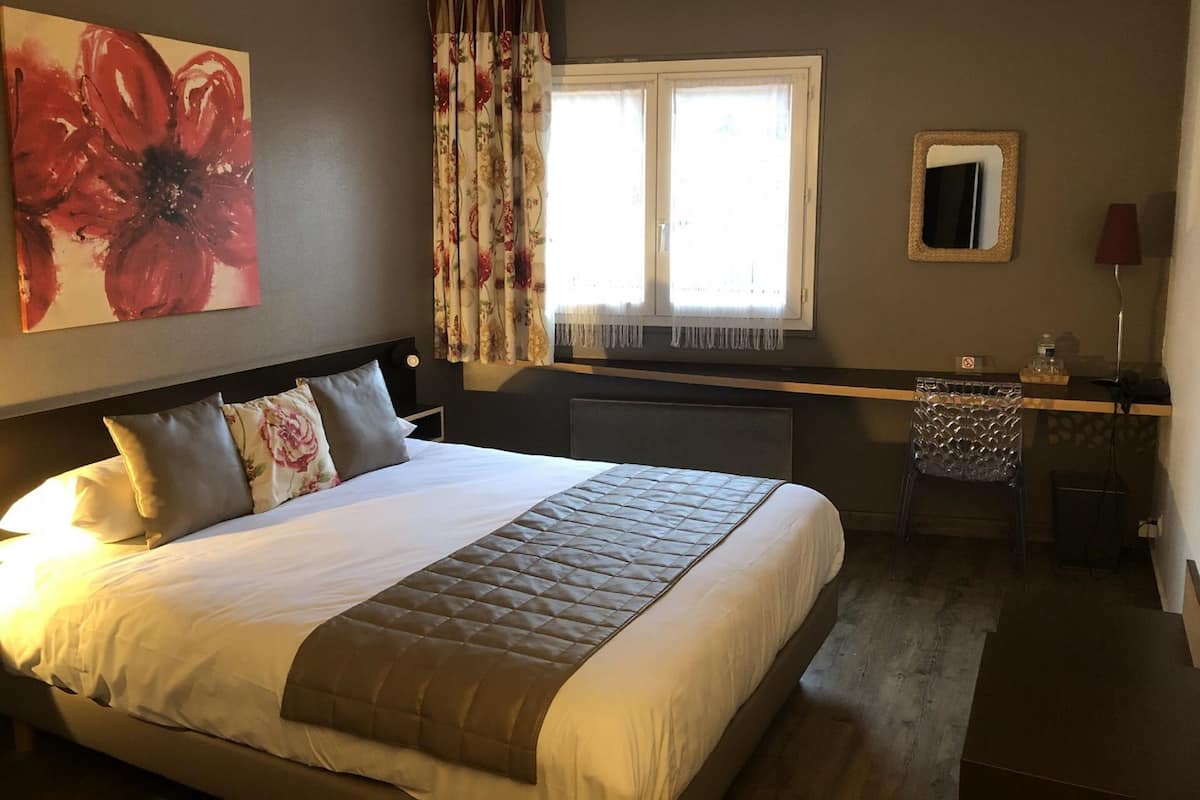 Una habitación de hotel con una cama grande, un escritorio con silla, una ventana con cortinas y un cuadro en la pared.