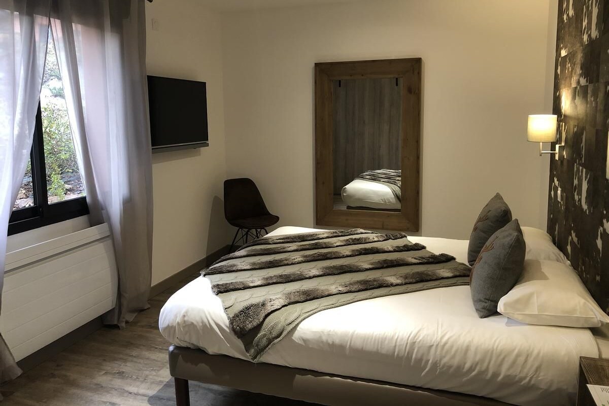 Un dormitorio con cama, una silla, televisor, ventana con cortinas y espejo adosado a la pared.
