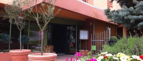 Fachada de hotel con toldo rojo, plantas en macetas y parterre de flores.