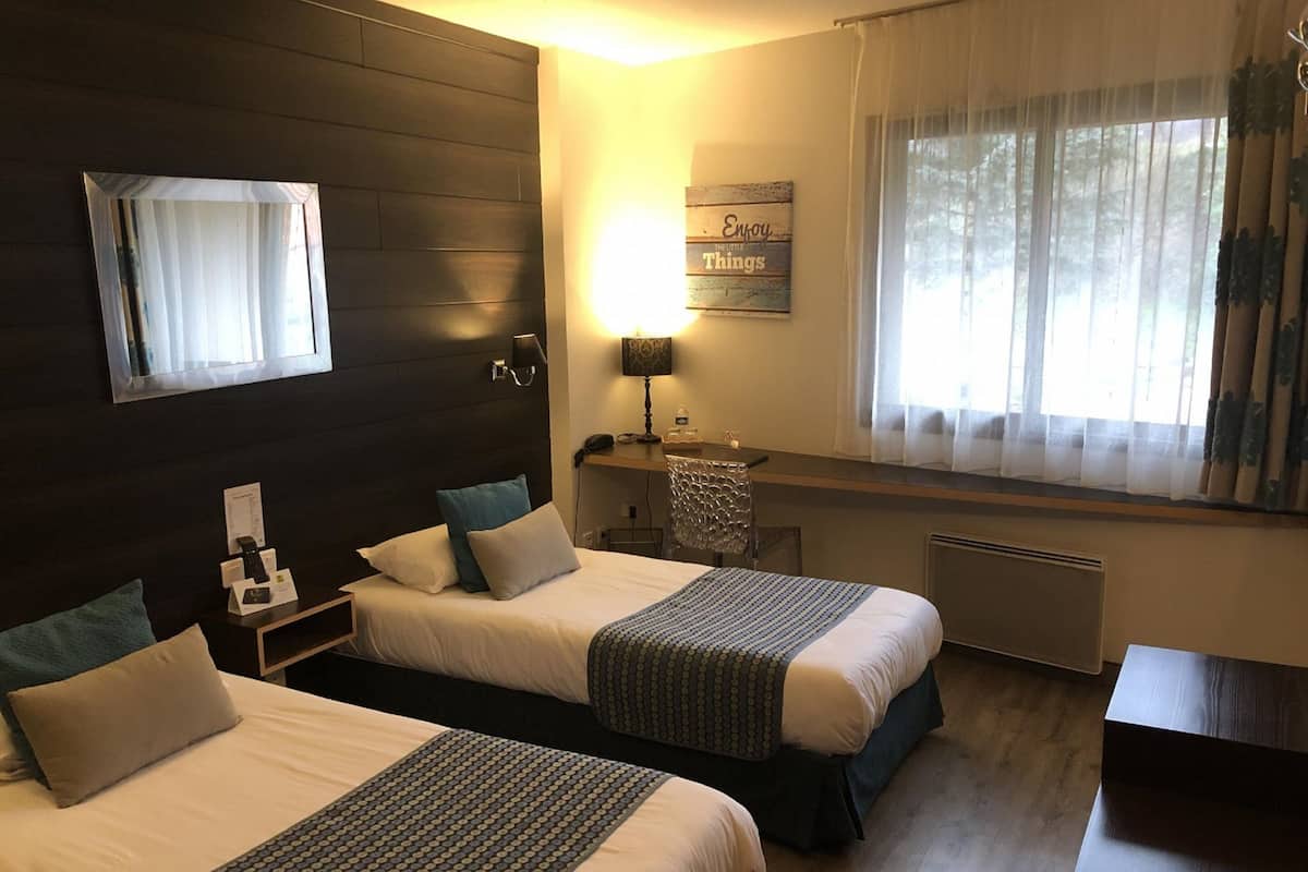 Habitación de hotel con dos camas, un escritorio, una silla, una lámpara y una ventana con cortinas.