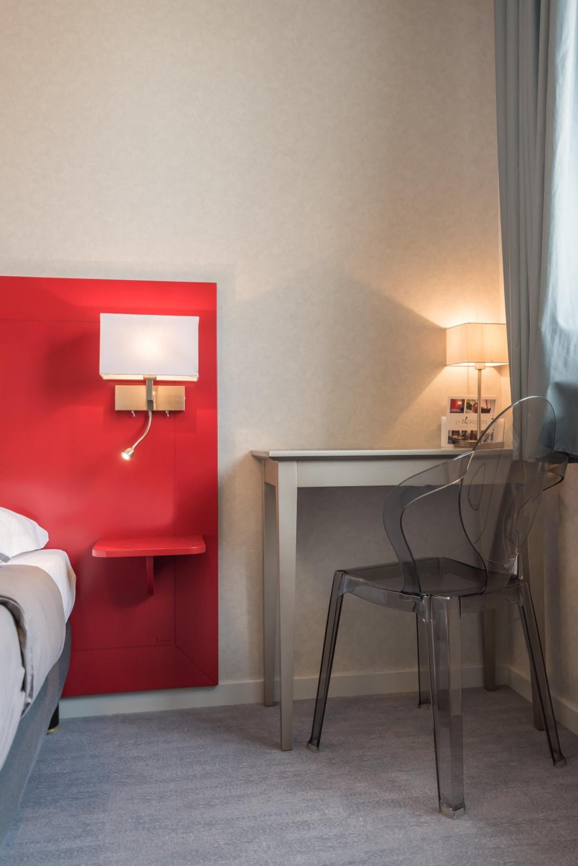 Ein Hotelzimmer mit roter Akzentwand, einem weißen Nachttisch, einem klaren Kunststoffstuhl, einem Schreibtisch mit Lampe und einem Bett.