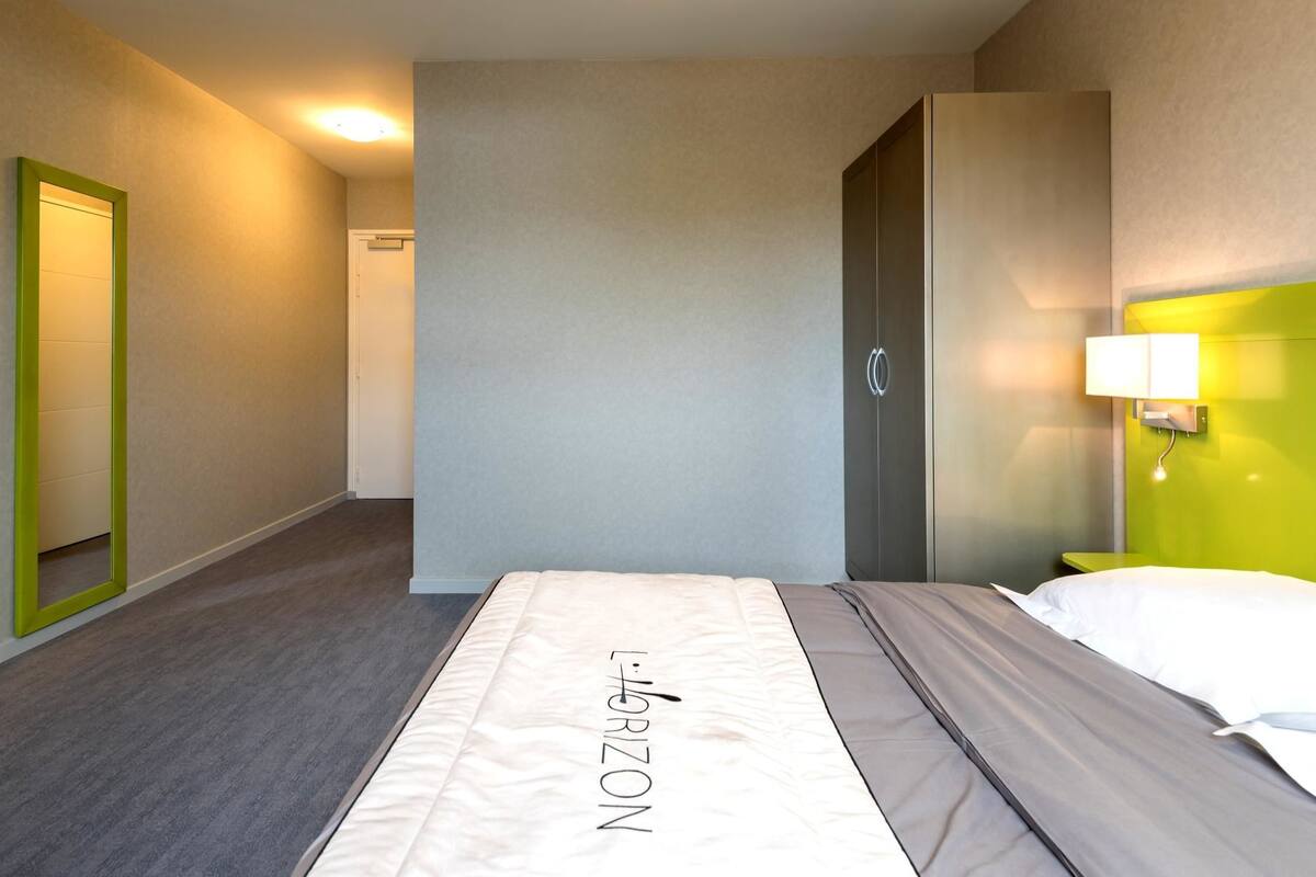 Une chambre d’hôtel avec un lit, un miroir encadré de vert, une porte et un luminaire fixé au mur.