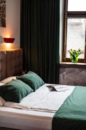 A neatly made bed with green bedding, a bedside lamp, and a window with a view of the city. - Aparthotel Maargick (Kraków)