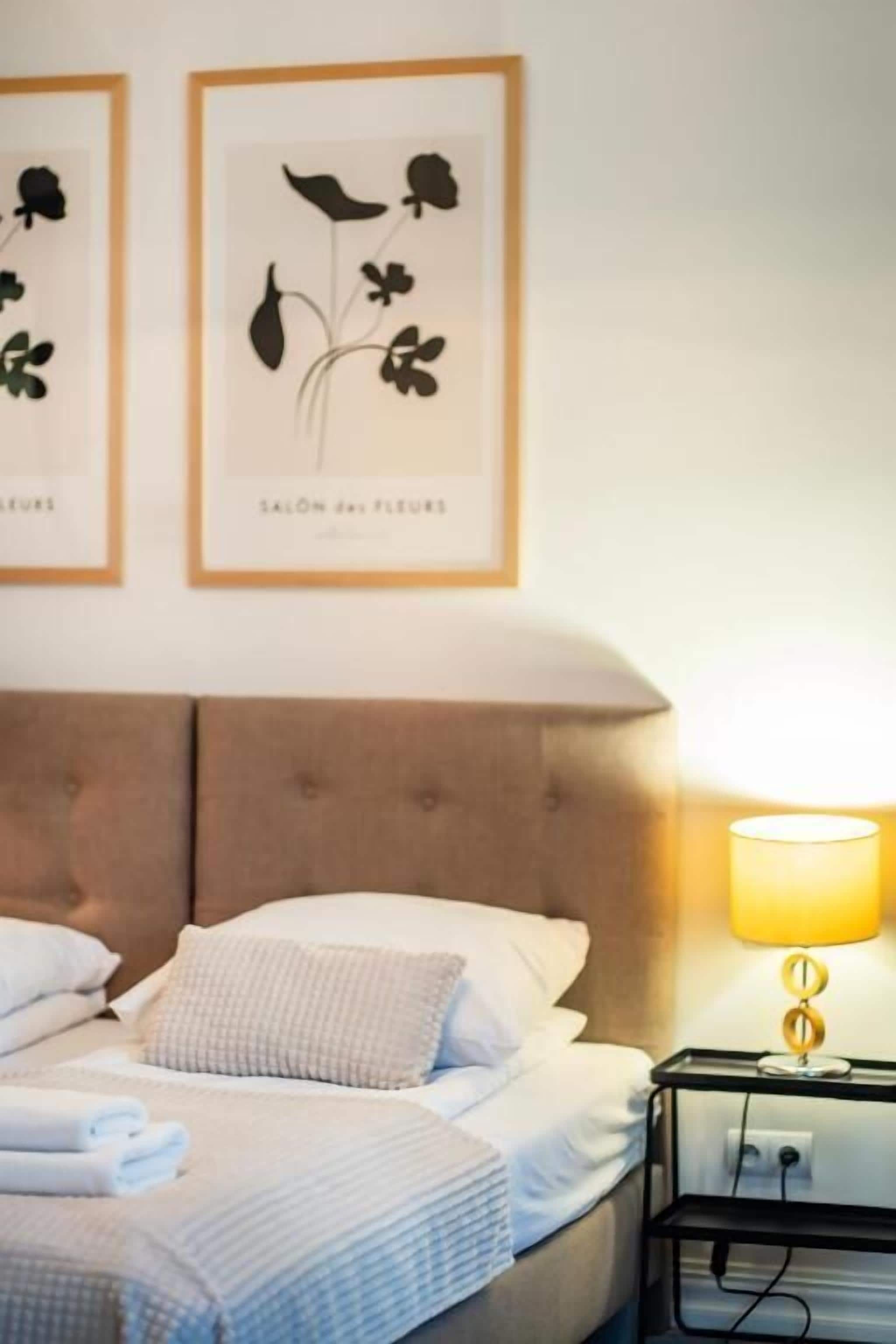 A hotel room with a bed, pillows, a bedside table with a lamp, and two framed botanical prints on the wall.