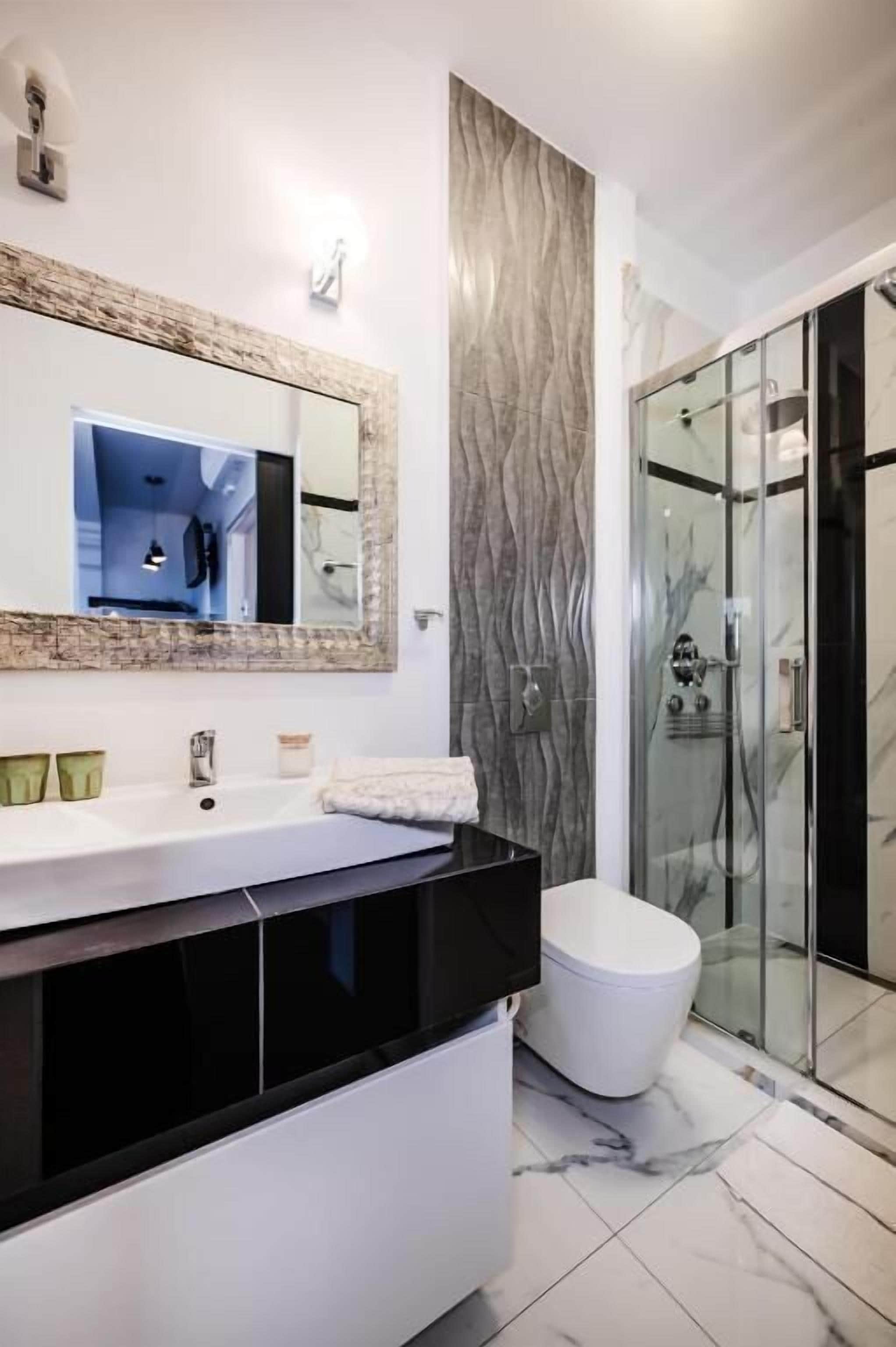 Un baño moderno con un espejo grande, lavamanos blanco, encimera negra y una cabina de ducha de vidrio.