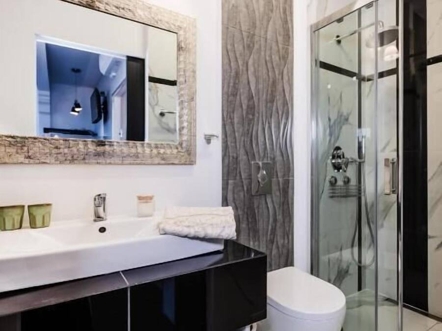 Un baño moderno con un espejo grande, lavamanos blanco, encimera negra y una cabina de ducha de vidrio.