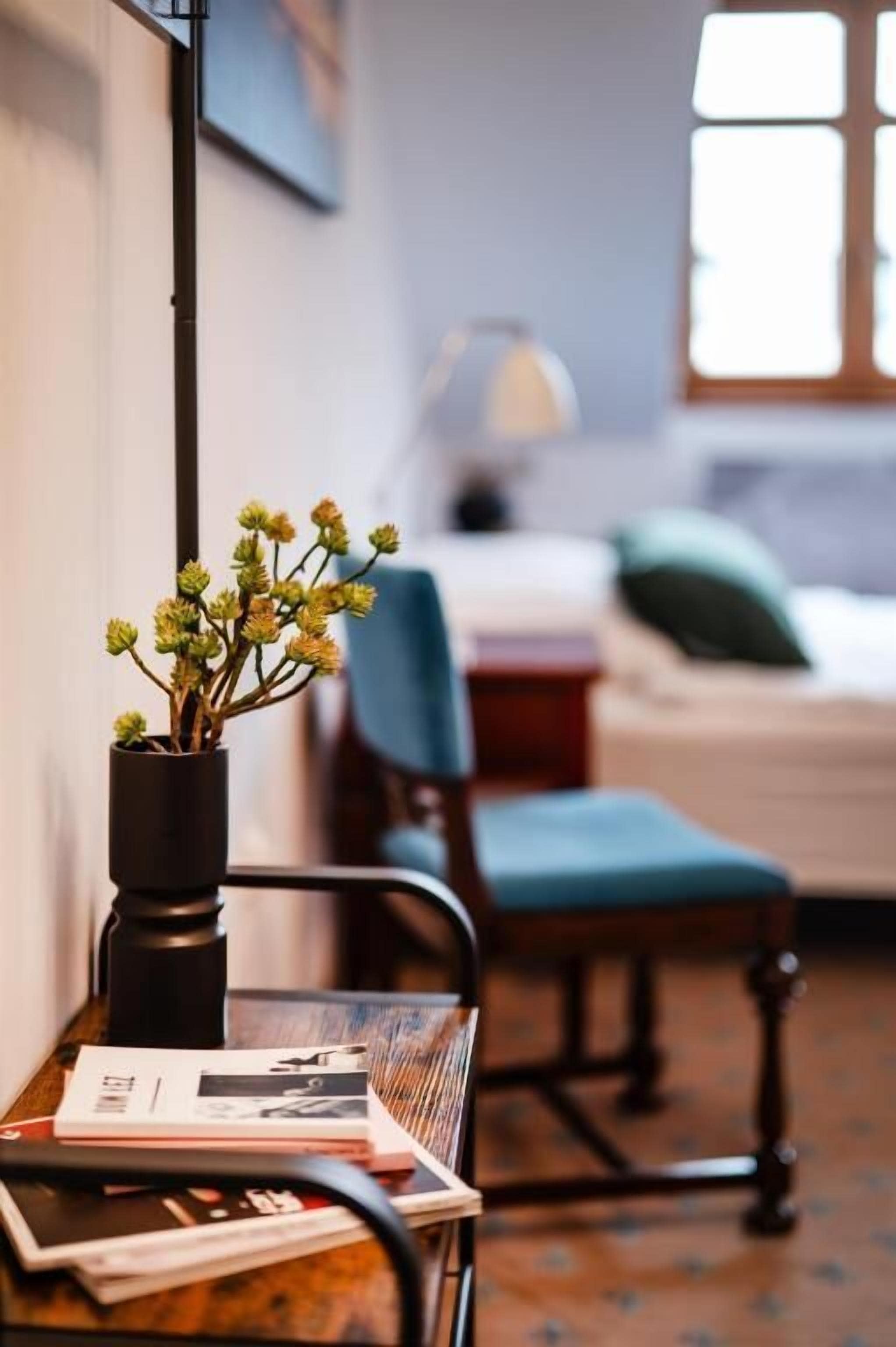 Una habitación con cama, una silla, una mesa con un florero y una lámpara.