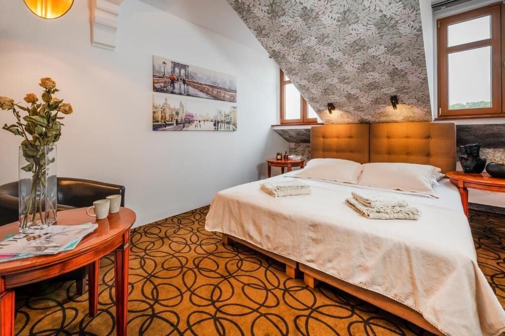 Une chambre d’hôtel avec un lit, une table ronde ornée d’un vase de fleurs, une chaise et un mur sur lequel est accrochée une peinture représentant un paysage urbain.