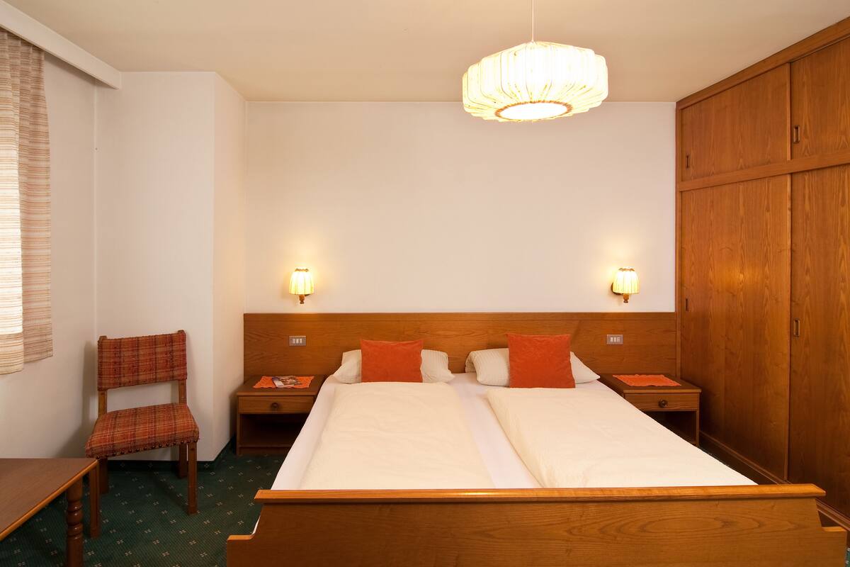 A hotel room with a wooden bed, two bedside tables, a chair, a wardrobe, and a chandelier.