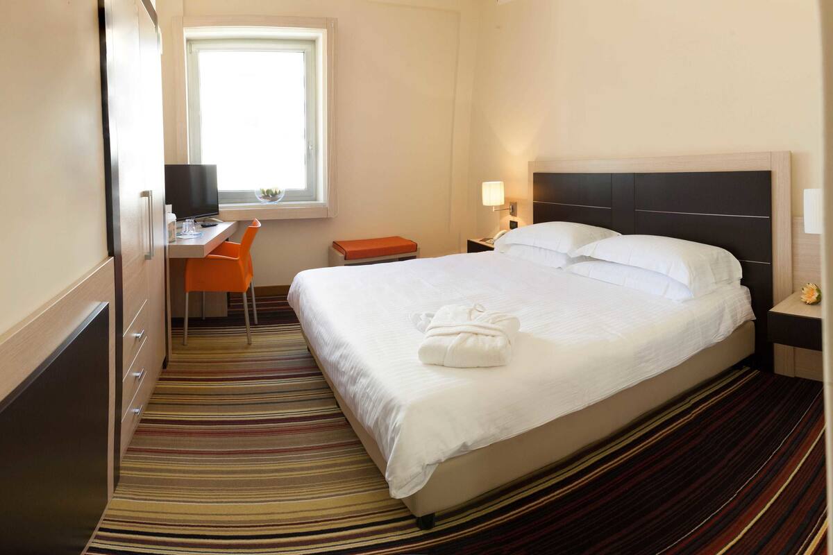 A hotel room with a bed, a desk with a chair, a TV, a window, and a nightstand with a lamp.