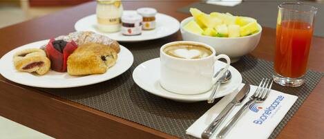 Desayuno con cruasanes, un bol de fruta, una taza de café y un vaso de jugo sobre una mesa.