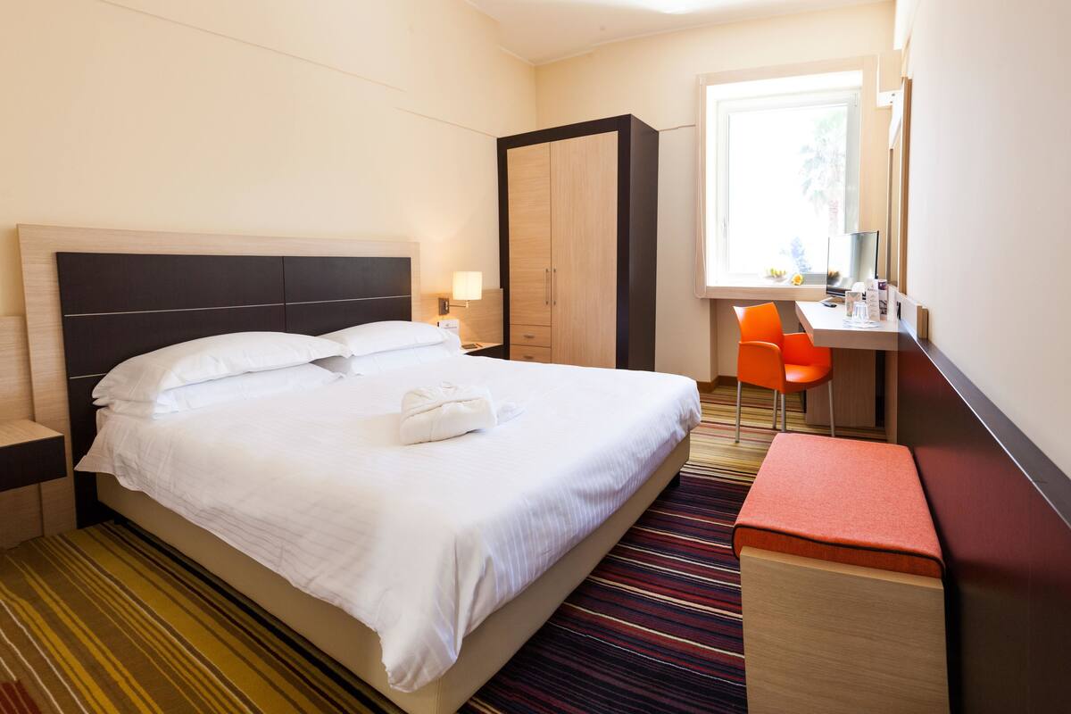 A hotel room with a bed, a desk, a chair, a wardrobe, and a window.
