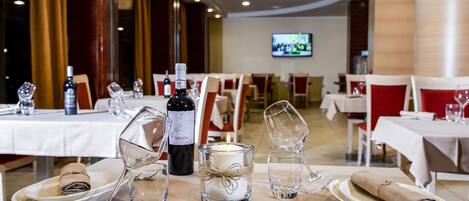 Mesa lista con platos, vasos, una vela y una botella de vino en un restaurante con sillas rojas y un televisor al fondo.