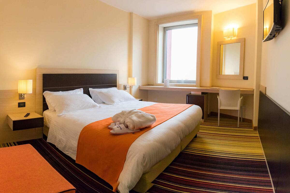 A hotel room with a bed, a desk, a chair, a window, and a mirror.