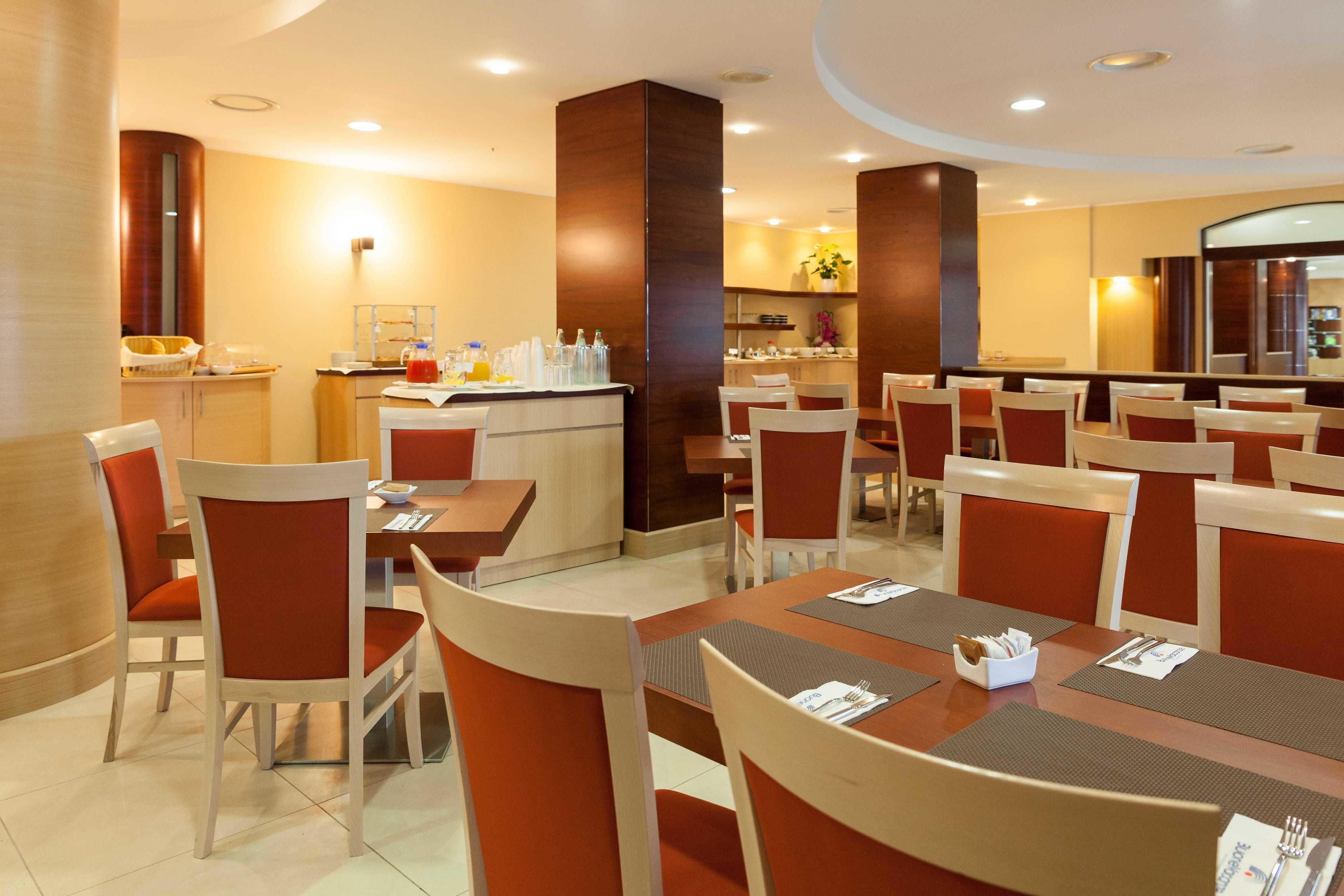 Une salle à manger d’hôtel avec de nombreuses tables et chaises, un comptoir central où sont servis les boissons, et un grand miroir qui agrandit visuellement l’espace.