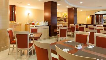 Une salle à manger d’hôtel avec de nombreuses tables et chaises, un comptoir central où sont servis les boissons, et un grand miroir qui agrandit visuellement l’espace.