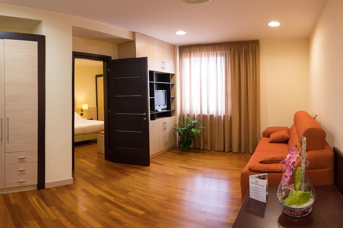 A hotel room with a wooden floor, a bed, a TV on a shelf, a sofa, and a sign with room amenities.