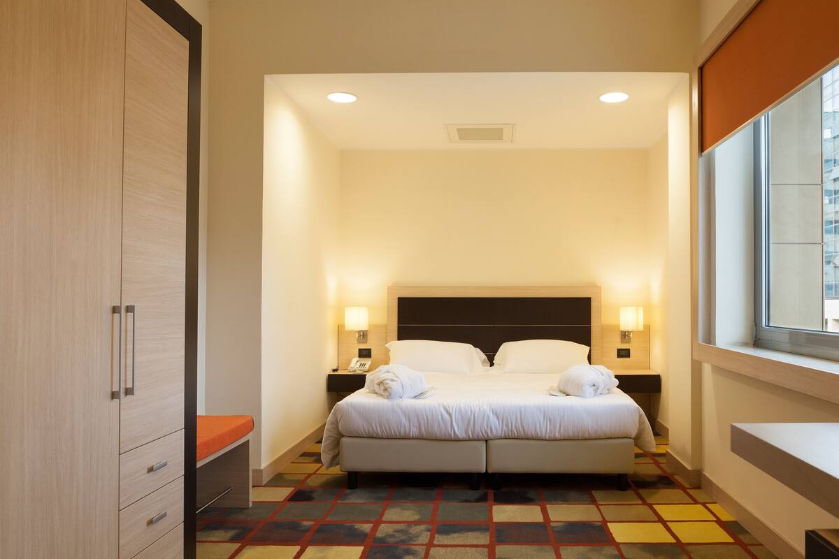A hotel room with a bed, bedside lamps, a window, and a wardrobe.