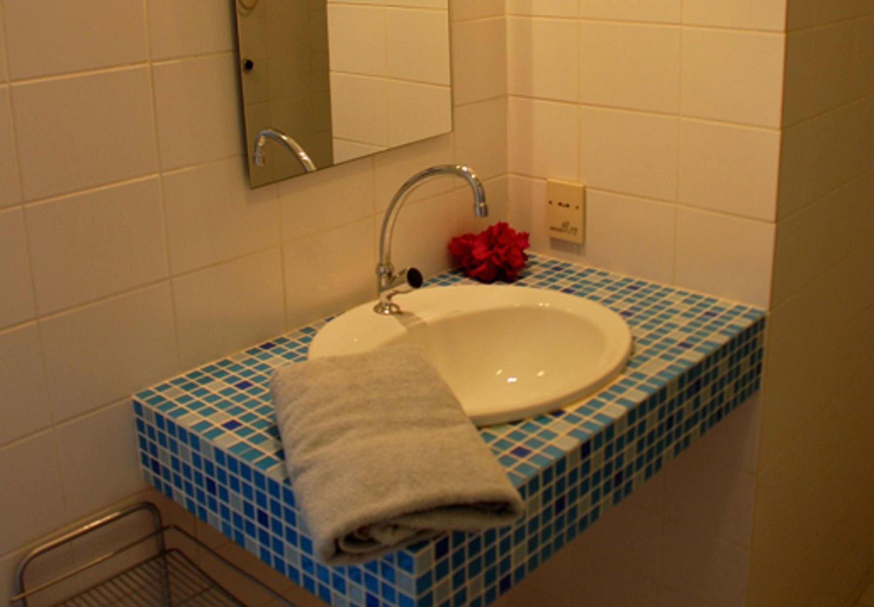Une salle de bain avec un lavabo blanc, des carreaux de mosaïque bleus, une serviette et une fleur rouge.