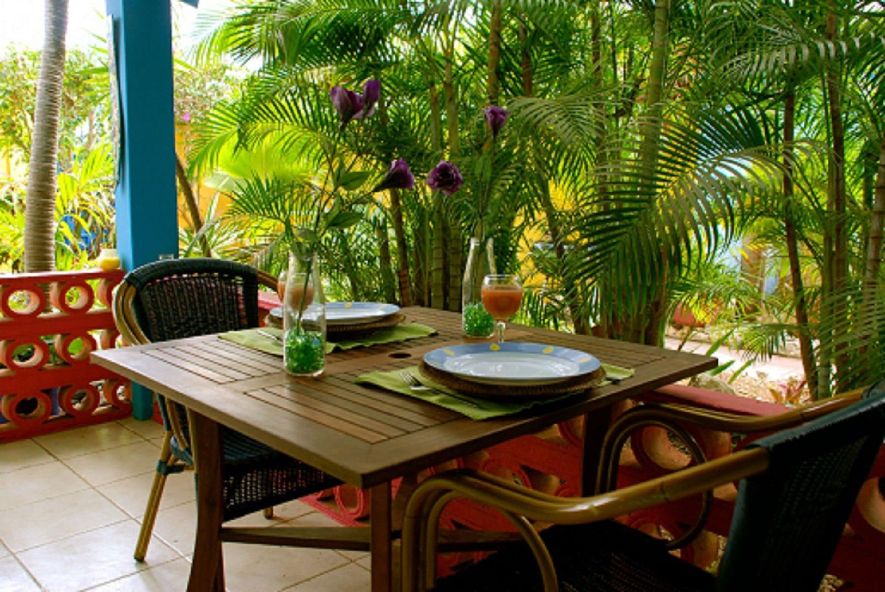 Mesa de jantar posta com pratos, copos e um vaso de flores, rodeada por cadeiras e um cenário tropical.