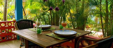 Mesa de jantar posta com pratos, copos e um vaso de flores, rodeada por cadeiras e um cenário tropical.