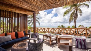 A covered outdoor seating area with wicker furniture, a blue sofa with orange cushions, and a view of the ocean and palm trees. - Pousada Vila Vagalume (Trairi)