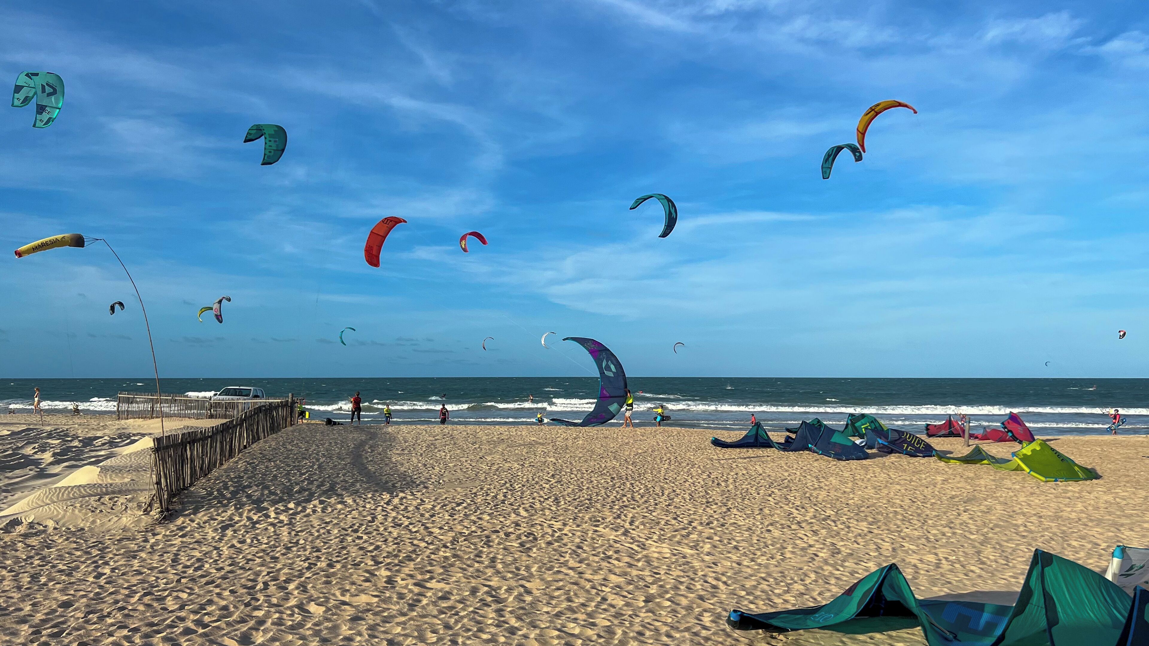 A beach with multiple kites flying in the sky, a wooden pier, and tents set up on the sand.