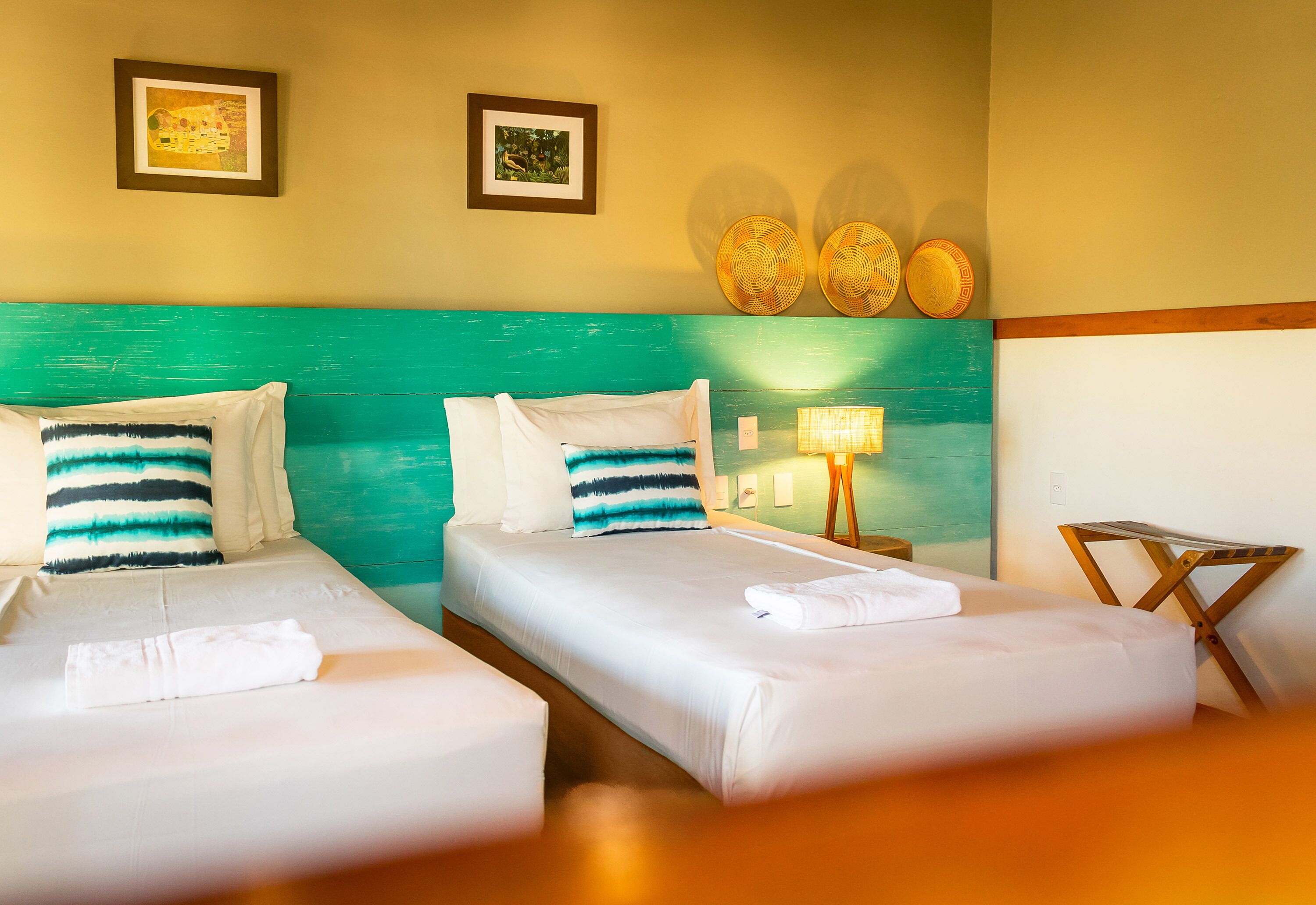 Quarto de hotel com duas camas, cada uma com roupa de cama branca e almofadas listradas, cabeceira verde-água e dois quadros na parede.
