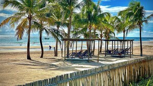 A beach with palm trees, a wooden fence, and lounge chairs under a wooden pergola. - Pousada Vila Vagalume (Trairi)