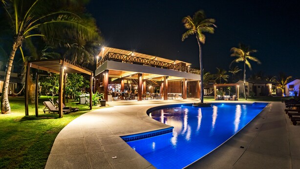 A nighttime swimming pool area with a covered seating area and palm trees. - Pousada Vila Vagalume (Trairi)