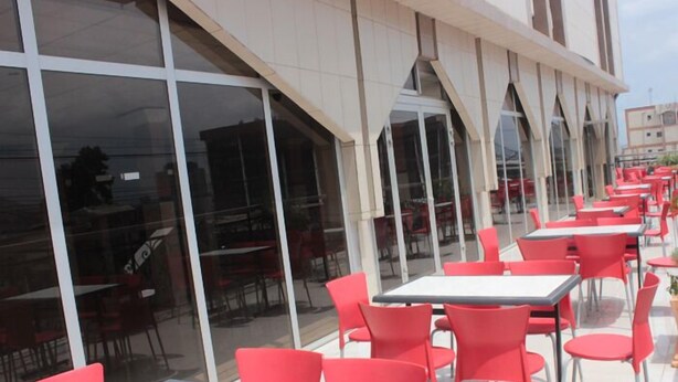 Un área de café al aire libre con sillas rojas y mesas blancas, ventanas amplias y una fachada de edificio moderna.