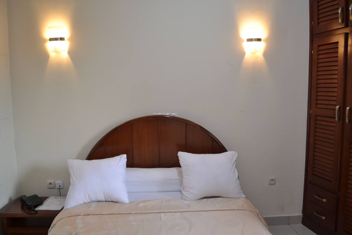A hotel room with a wooden headboard, two bedside lamps, a bedside phone, and a neatly made bed with white bedding.