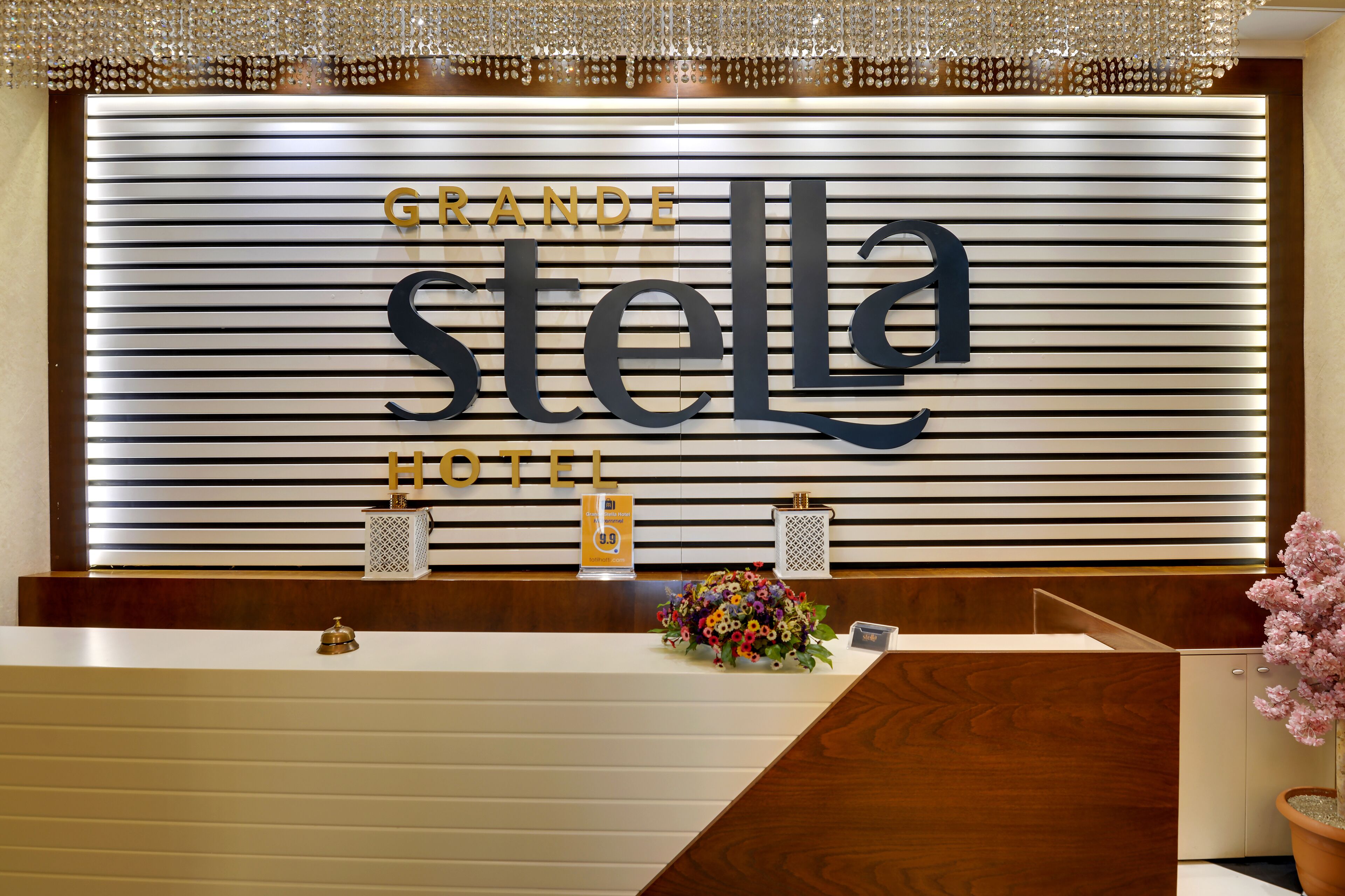 Una recepción de hotel con diseño moderno, que incluye un gran letrero que dice “GRANDE STELLA HOTEL” y un área de recepción con mostrador de madera, un arreglo floral y una planta en maceta.