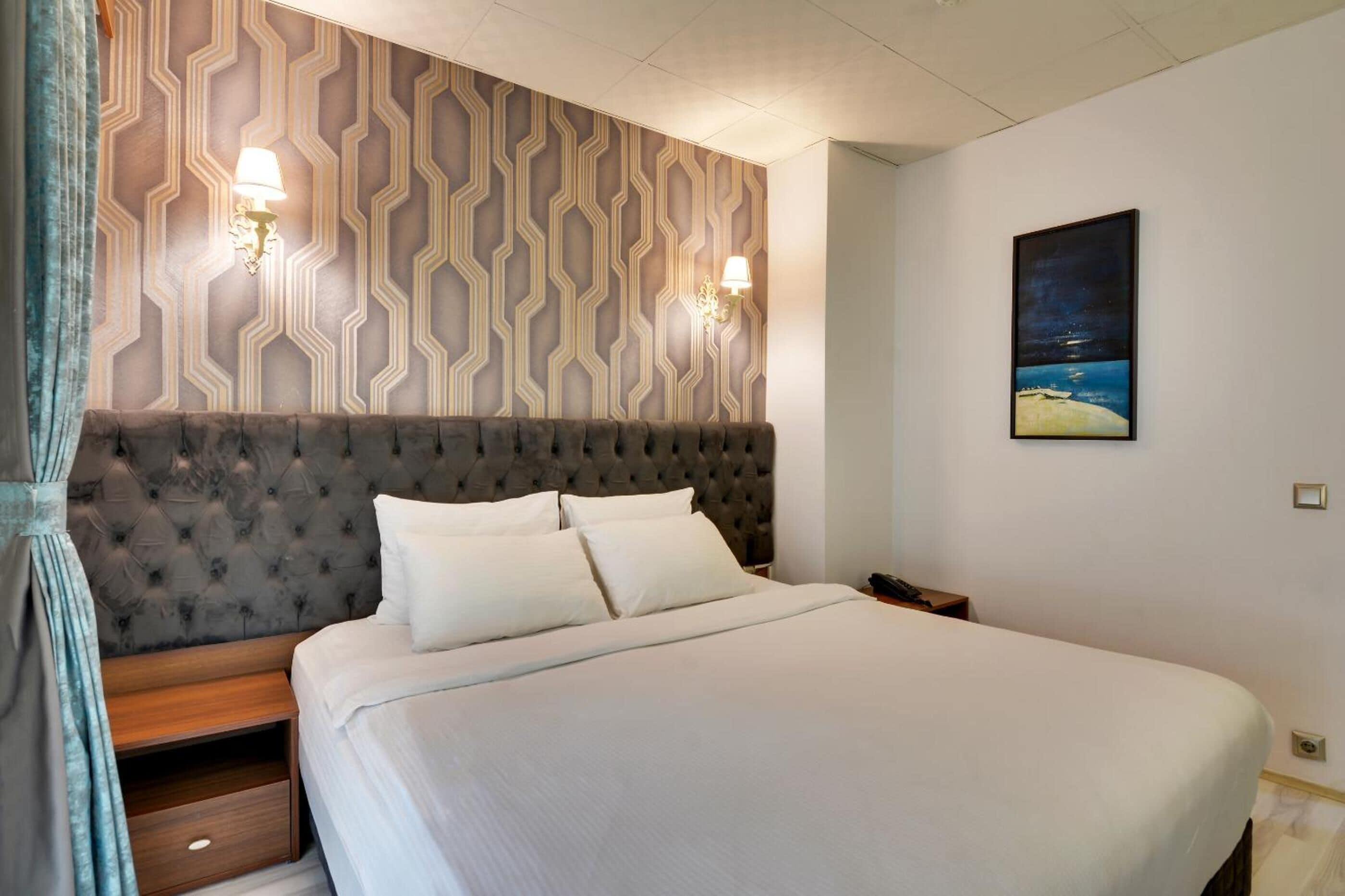 Una cama bien hecha con un cabecero capitonado, dos lámparas de pared y un cuadro enmarcado con una escena de playa.