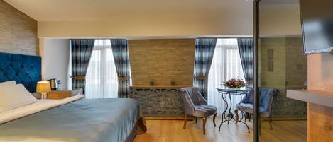 Quarto de hotel moderno com uma cama grande, área de estar com duas cadeiras, uma pequena mesa e uma televisão.