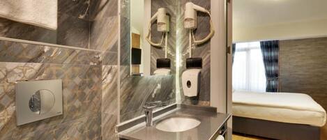 A modern bathroom with a marble backsplash, a sink, a mirror, a towel dispenser, and a bed in the adjacent room.