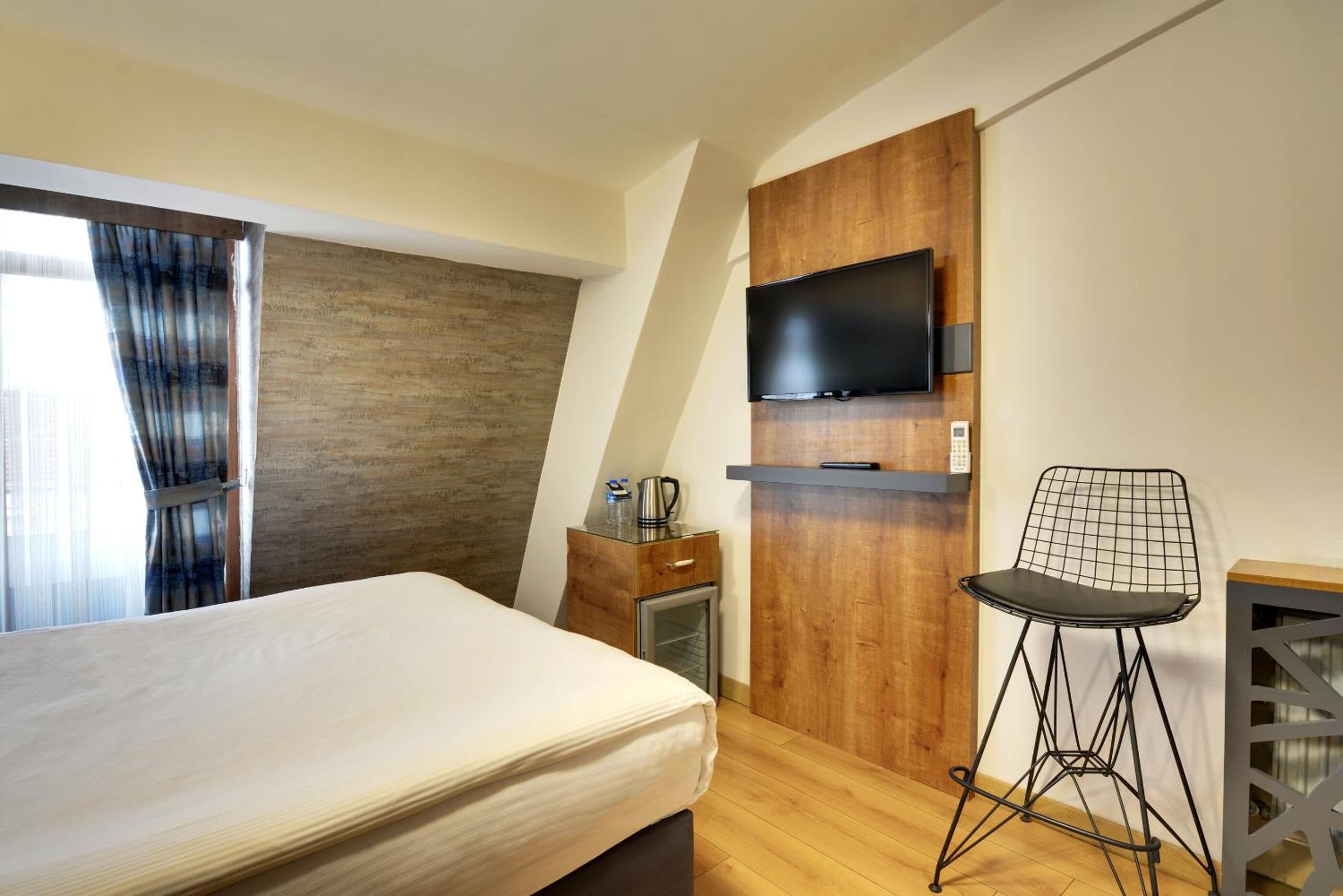 A bedroom with a bed, a TV mounted on a wooden wall, a small table with a kettle, and a chair.